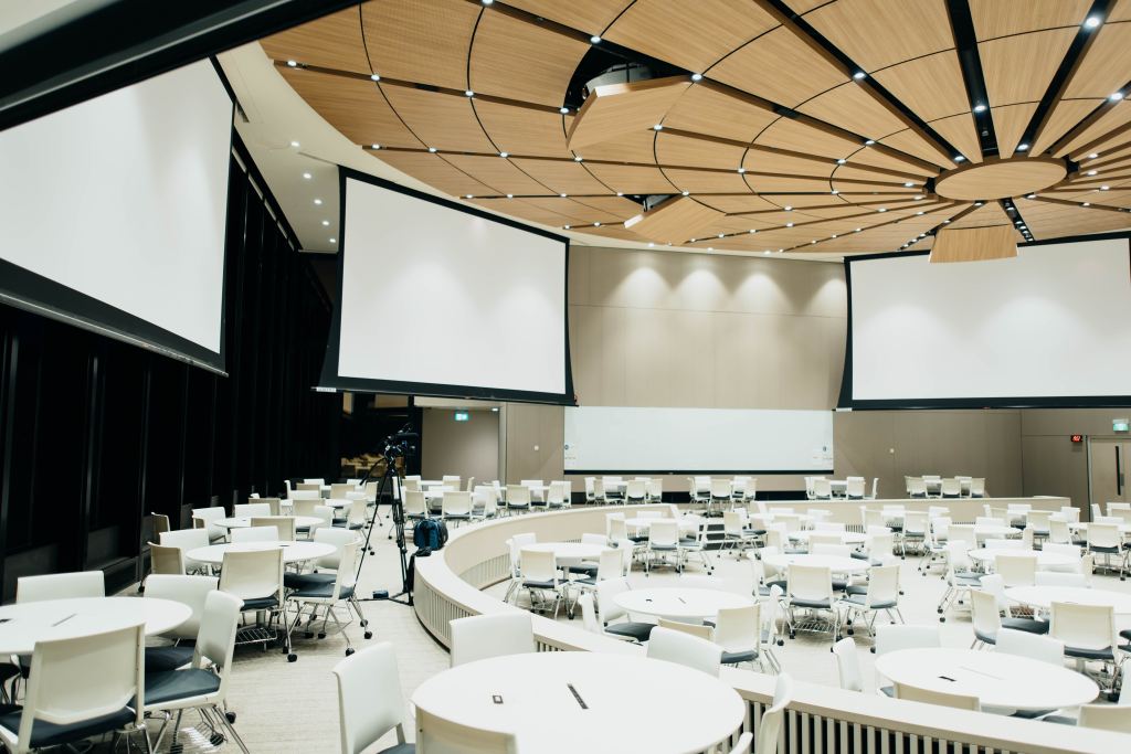 Chuttersnap on Unsplash Large round conference room with raised outer ring. The room is filled with white round tables, white and gray rolling chairs. There are large drop down projection screens hung around the room.