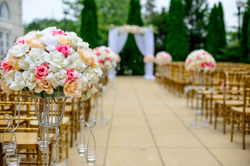 Shardayyy on Unsplash Beautiful outdoor wedding venue with tiled floor, gold Chivari chairs, peach, white, and pink flowers and an arch in front of green trees.