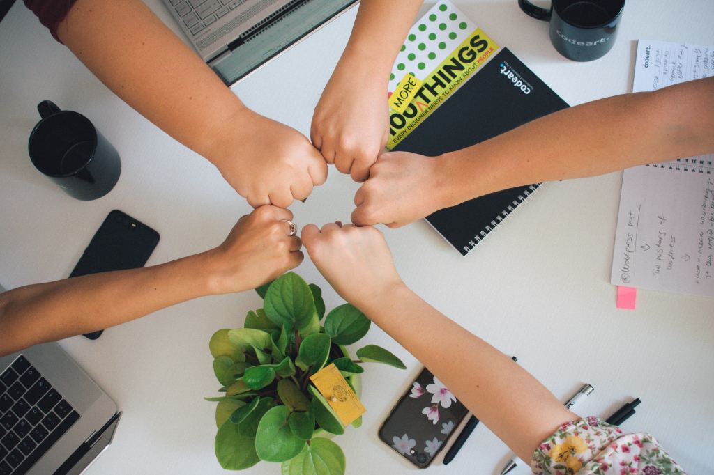 A circle made of 5 people's fists above an office table with work and computers beneath