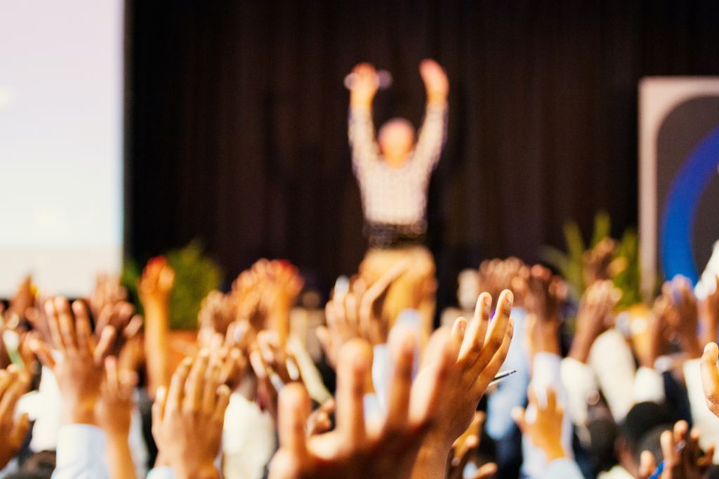A group of people raise both of their hands up high mimicking a man on stage.