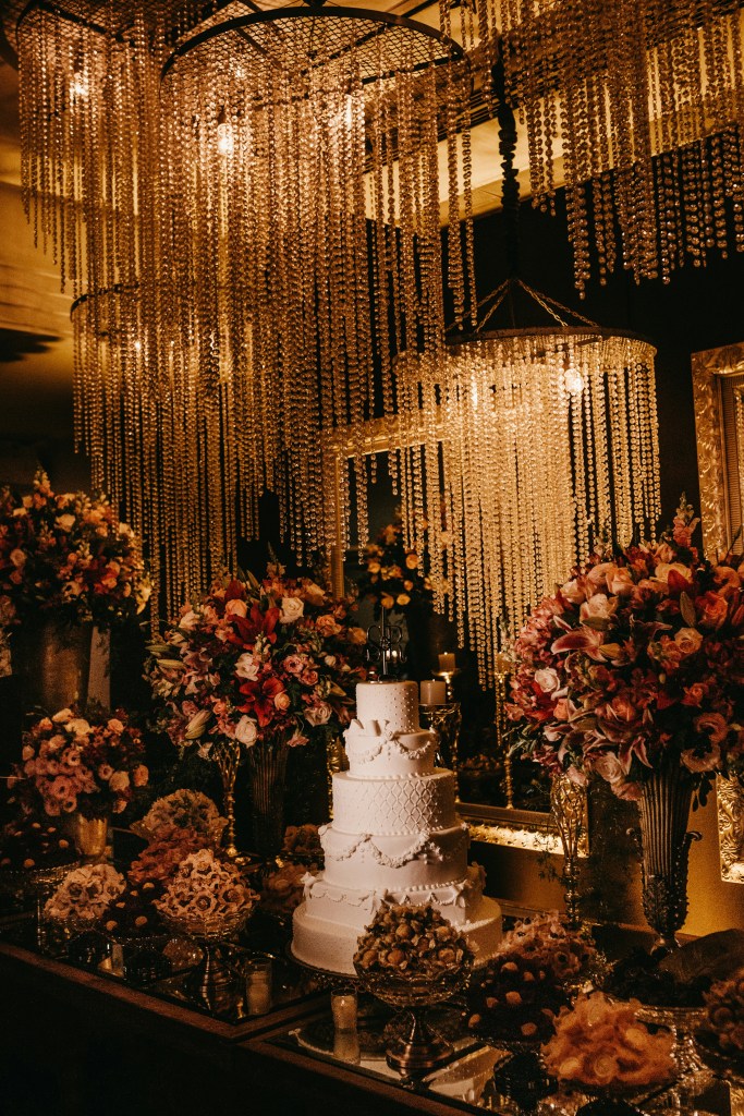 Extravagant dripping pearls chandeliers placed closely together over an all white decorative 5 tier wedding cake surrounded by huge mutlicolored red and pink florals in gold vases and golden bowls of other kinds of fancy desserts