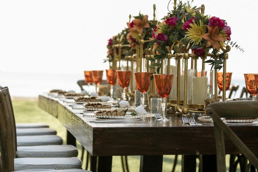 Tall raised florals on gold stand with clear tablesettings and rust colored water glasses on a bare table oustide